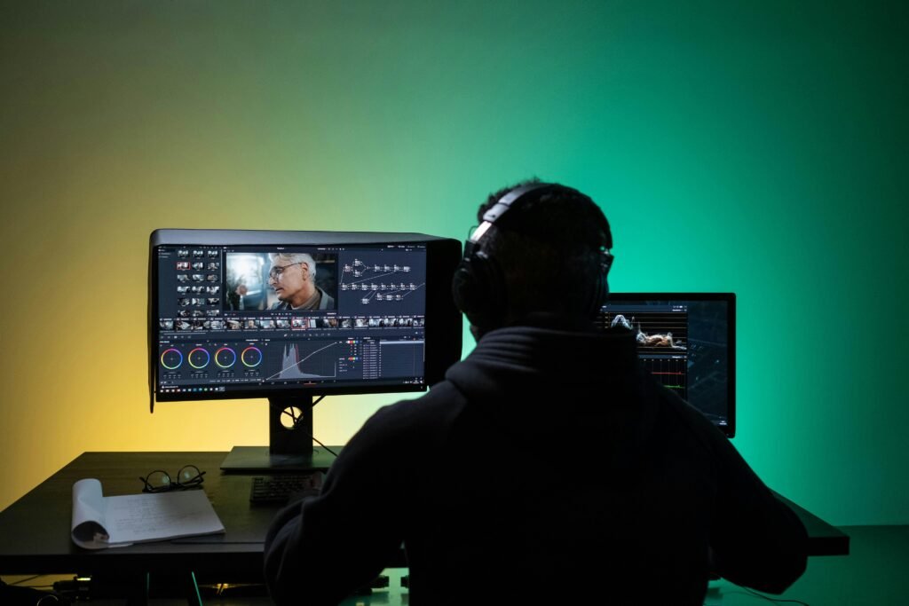 A person wearing headphones works on video editing at a desk with dual monitors displaying a film editing software interface, featuring color grading and various video clips.